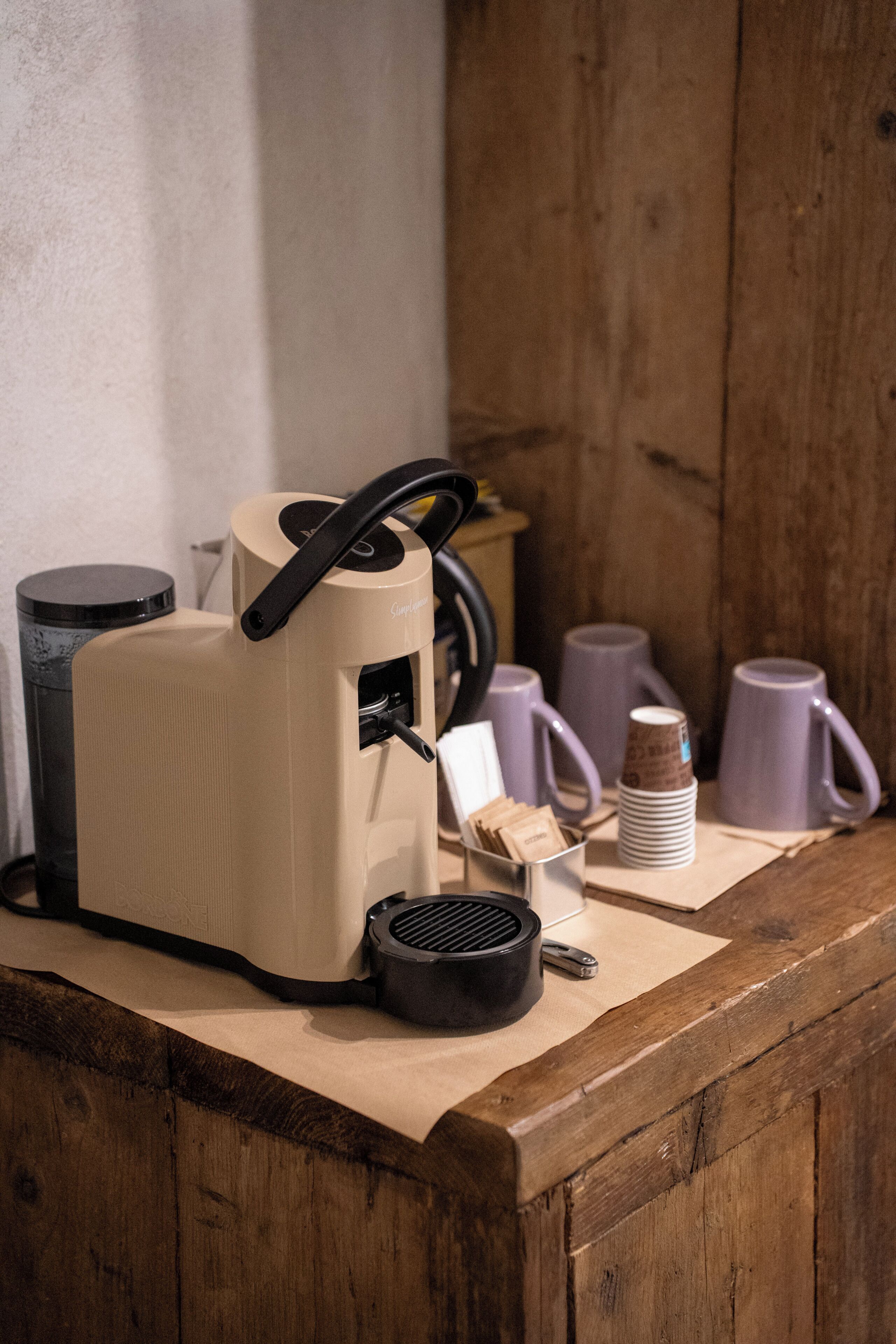 Máquina de café sobre bancada de madeira, com xícaras empilhadas e um cesto de lixo.
