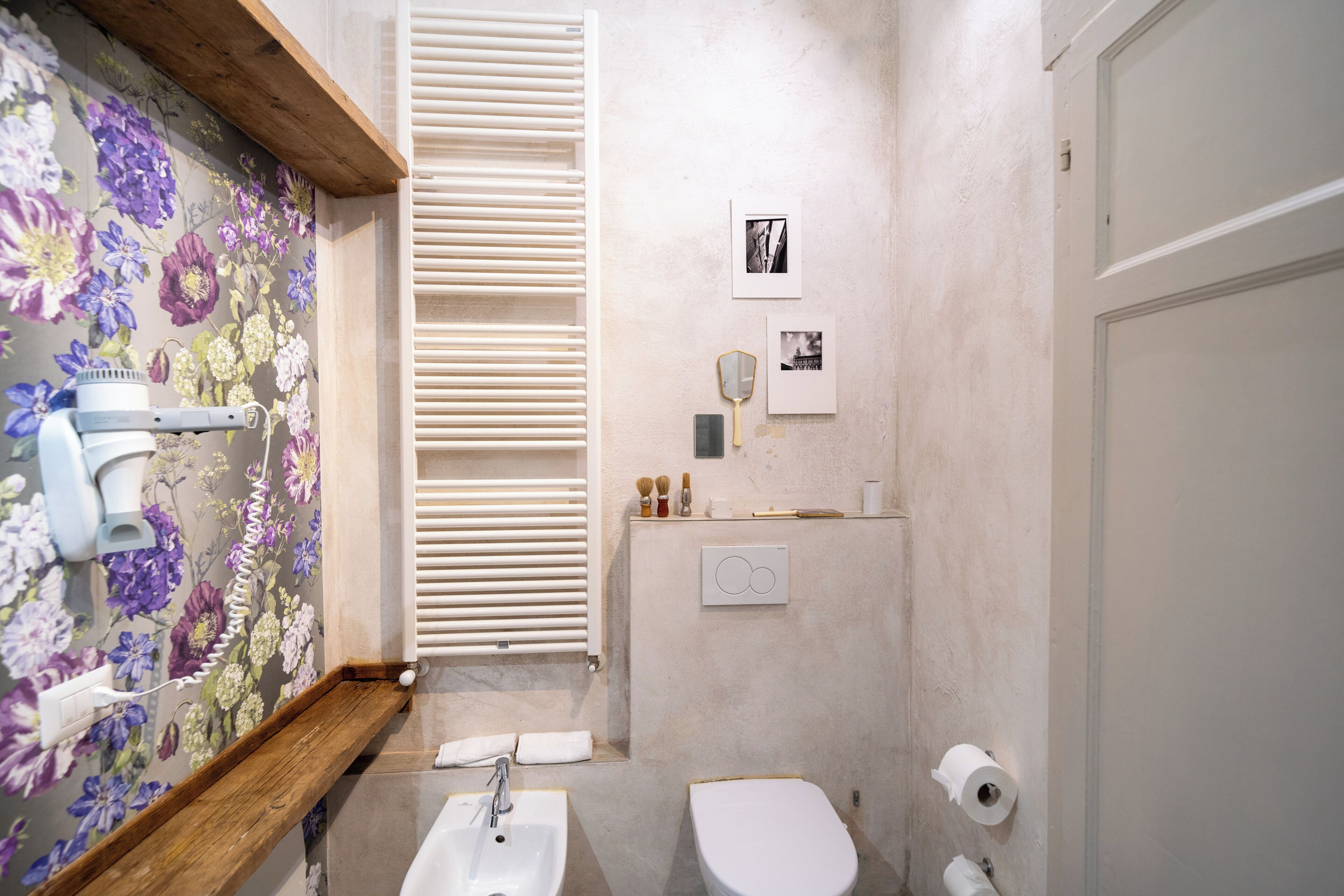 A bathroom with a floral wallpaper, a wooden shelf, a white sink, a toilet, and a towel rack.