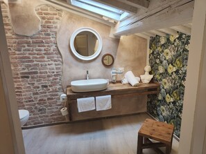A bathroom with a brick wall, a wooden vanity, a round mirror, a white sink, a wooden stool, and a floral wallpaper.