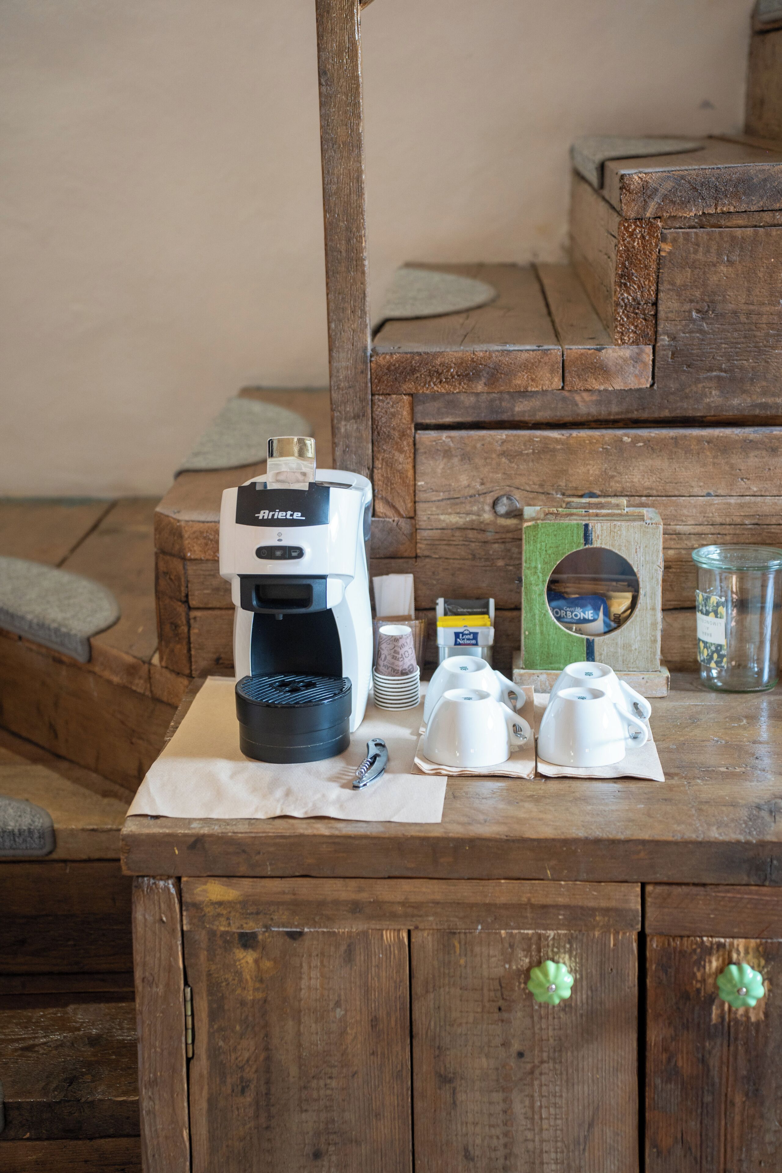 A wooden table with a coffee maker, cups, and a jar.