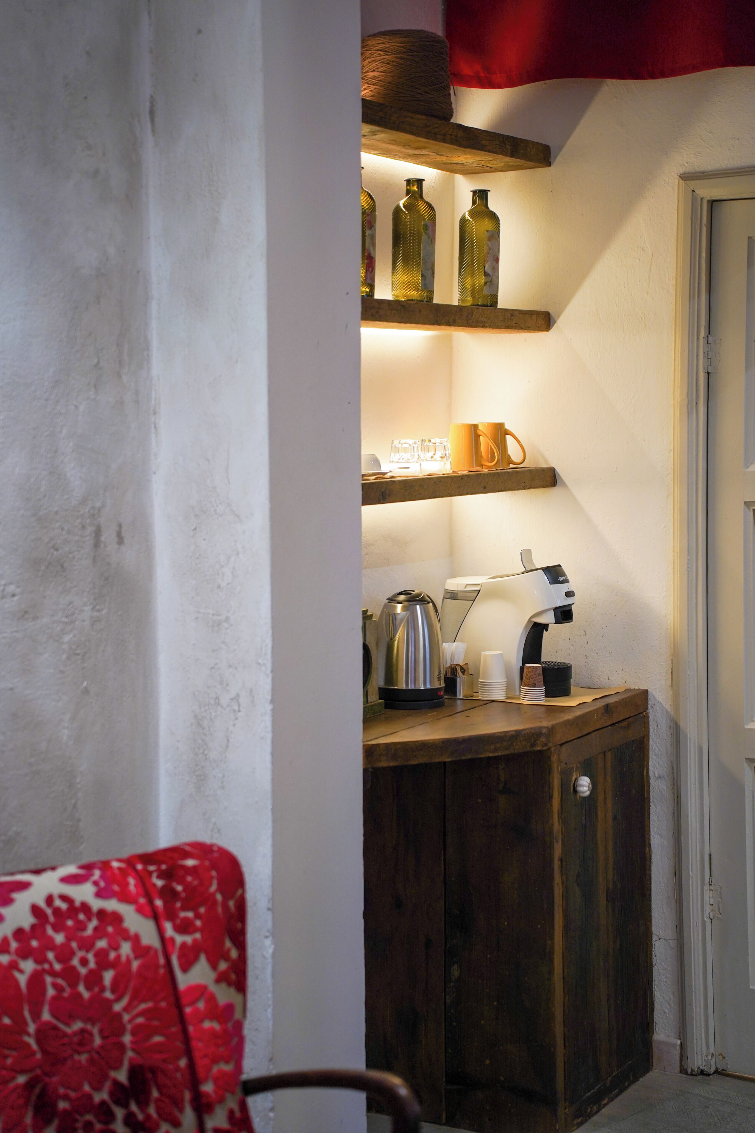 A corner of a room with a wooden cabinet, a coffee machine, and a shelf with bottles and a cup.