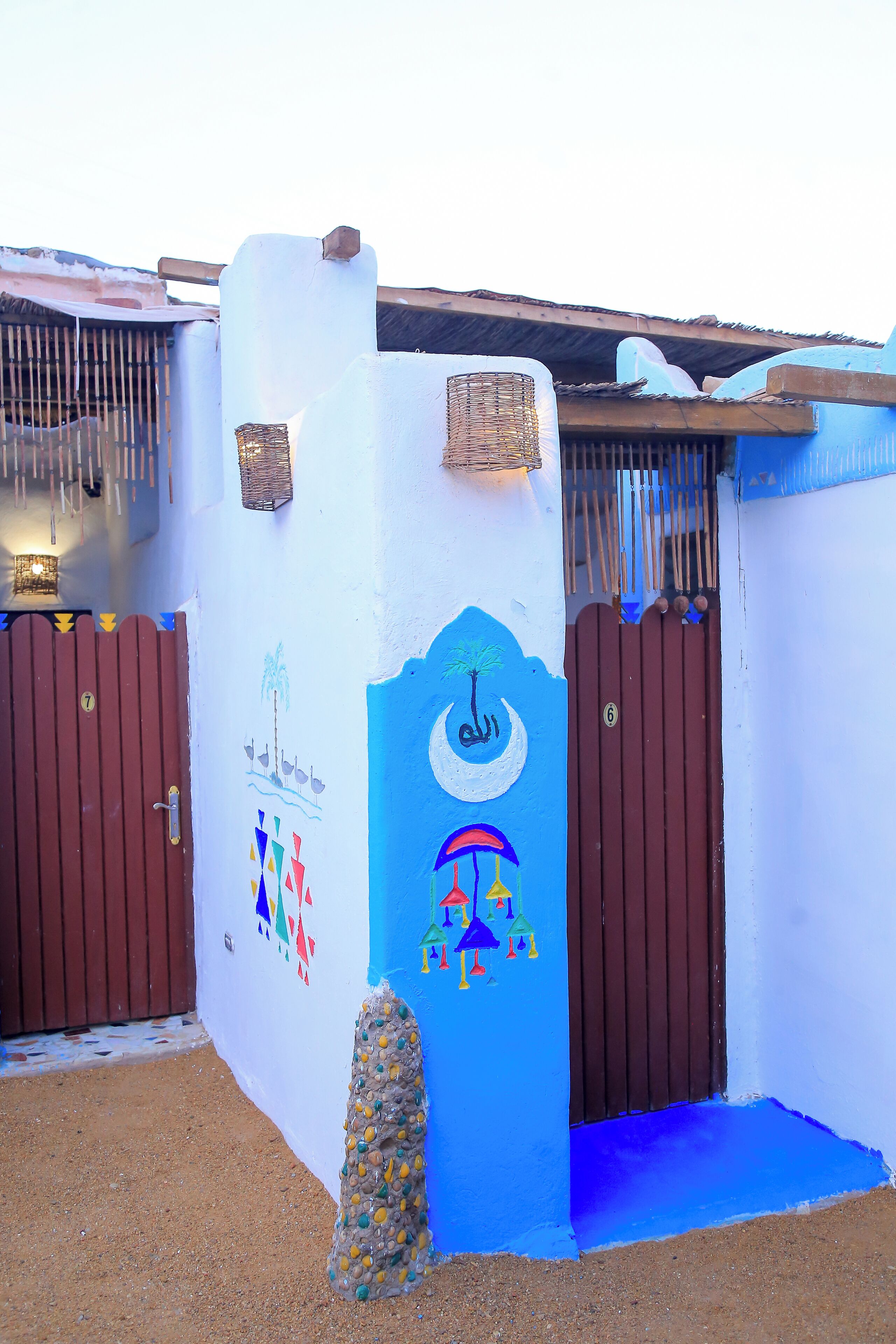 Un edificio con fachada azul y blanca, que presenta una pared decorativa con motivos culturales, dos portones marrones y una pequeña construcción de piedra.