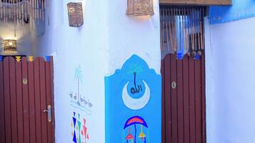 Un edificio con fachada azul y blanca, que presenta una pared decorativa con motivos culturales, dos portones marrones y una pequeña construcción de piedra.