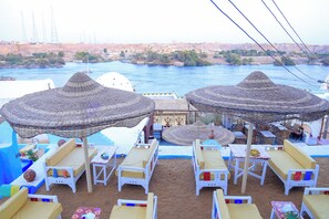 A beachside lounge with white lounge chairs and thatched umbrellas, overlooking a river with boats and a distant cityscape. - Nubian Palace Eco Lodge - A Nubian Home Since 1959 (Aswan)