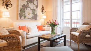 Un salón con un sofá blanco, dos sillones de mimbre, una mesa baja con libros, un jarrón con flores y un cuadro floral grande en la pared.