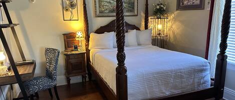 A four-poster bed with a wooden headboard, a nightstand with a lamp, a chair, and framed artwork on the walls.