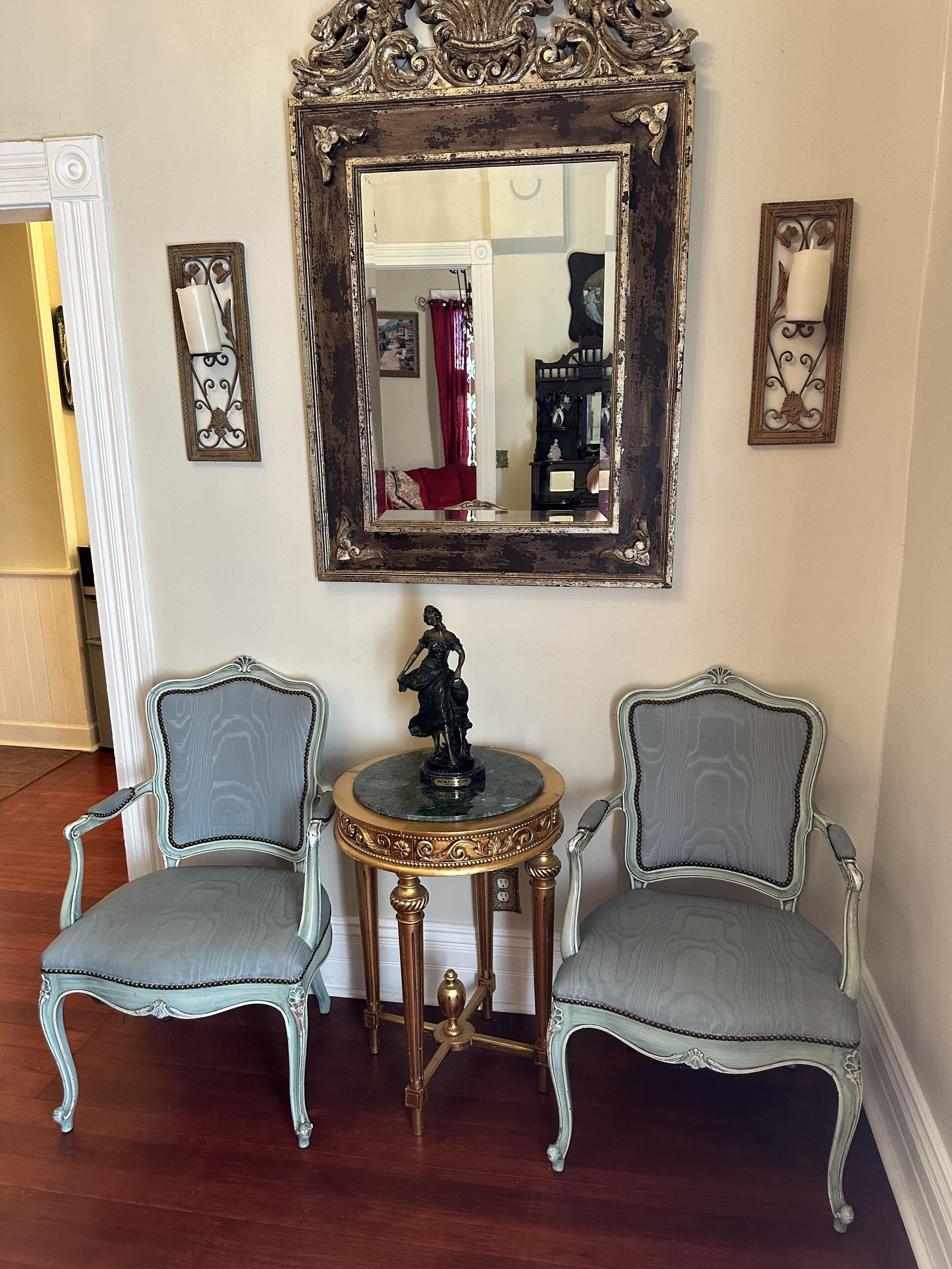 A room with two ornate chairs, a small table with a statue, and a large mirror with an ornate frame.