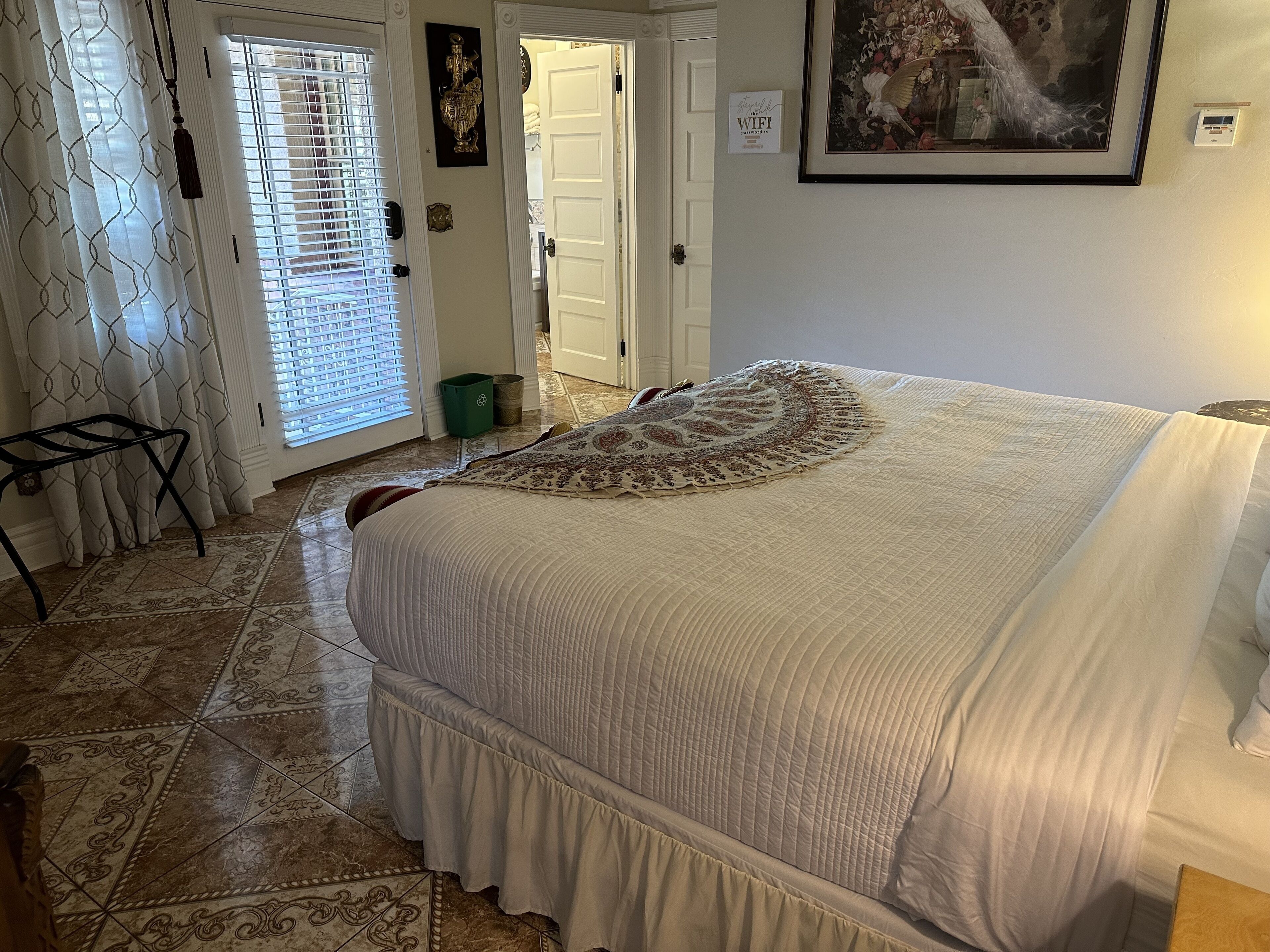 A bedroom with a large bed, a door with a sign, a framed picture on the wall, and a tiled floor.