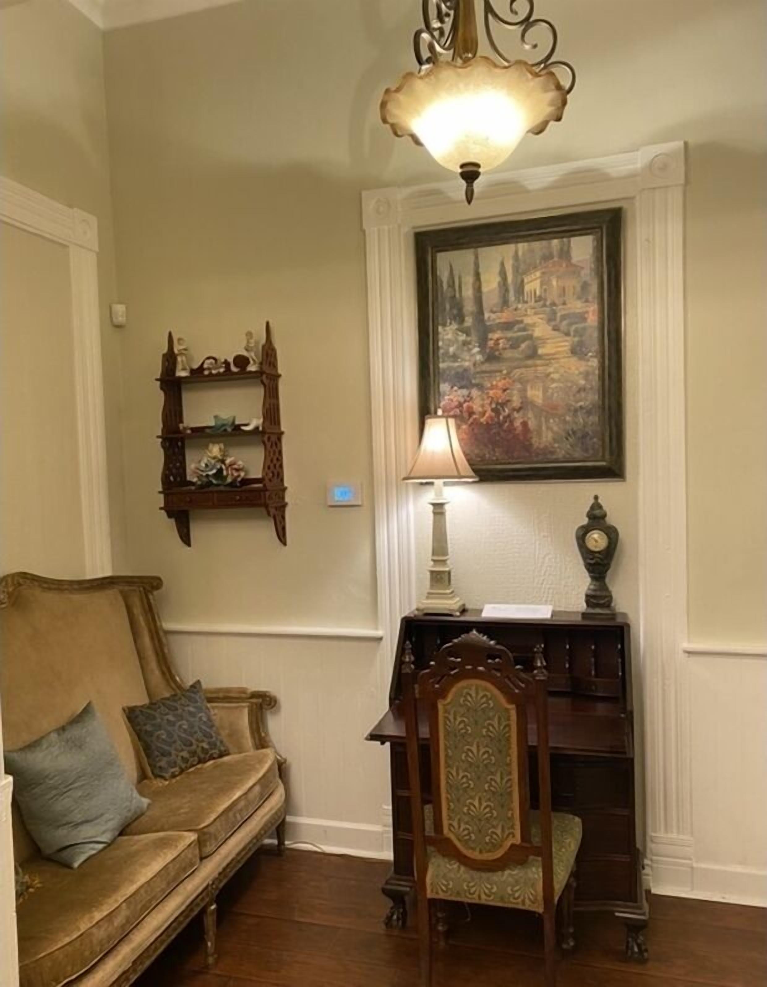 A room with a beige wall, a wooden chair with a green cushion, a wooden desk with a lamp, and a painting above the desk.