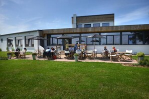 An outdoor dining area with tables and chairs, a building with large windows, and a grassy lawn. - l'Aérodrome de la Baie de Somme (Buigny-Saint-Maclou)