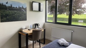 A hotel room with a bed, a desk with a chair, a window with a view of greenery, a radiator, and a painting on the wall. - l'Aérodrome de la Baie de Somme (Buigny-Saint-Maclou)