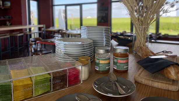 A dining area with a view of the outdoors, featuring a table with stacked plates, jars of condiments, and a vase with dried plants. - l'Aérodrome de la Baie de Somme (Buigny-Saint-Maclou)
