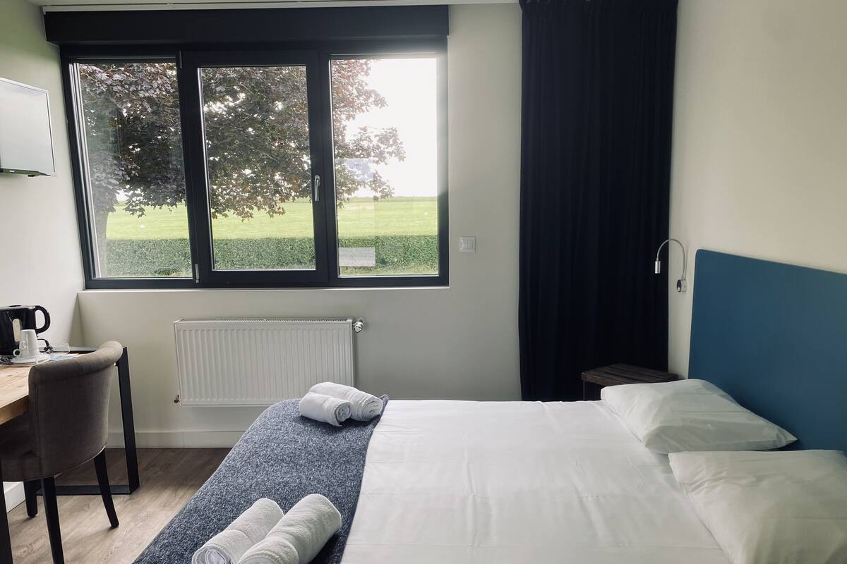 Ein Hotelzimmer mit Bett, Schreibtisch mit Kaffeemaschine, Stuhl und Fenster mit Blick auf eine grüne Landschaft.