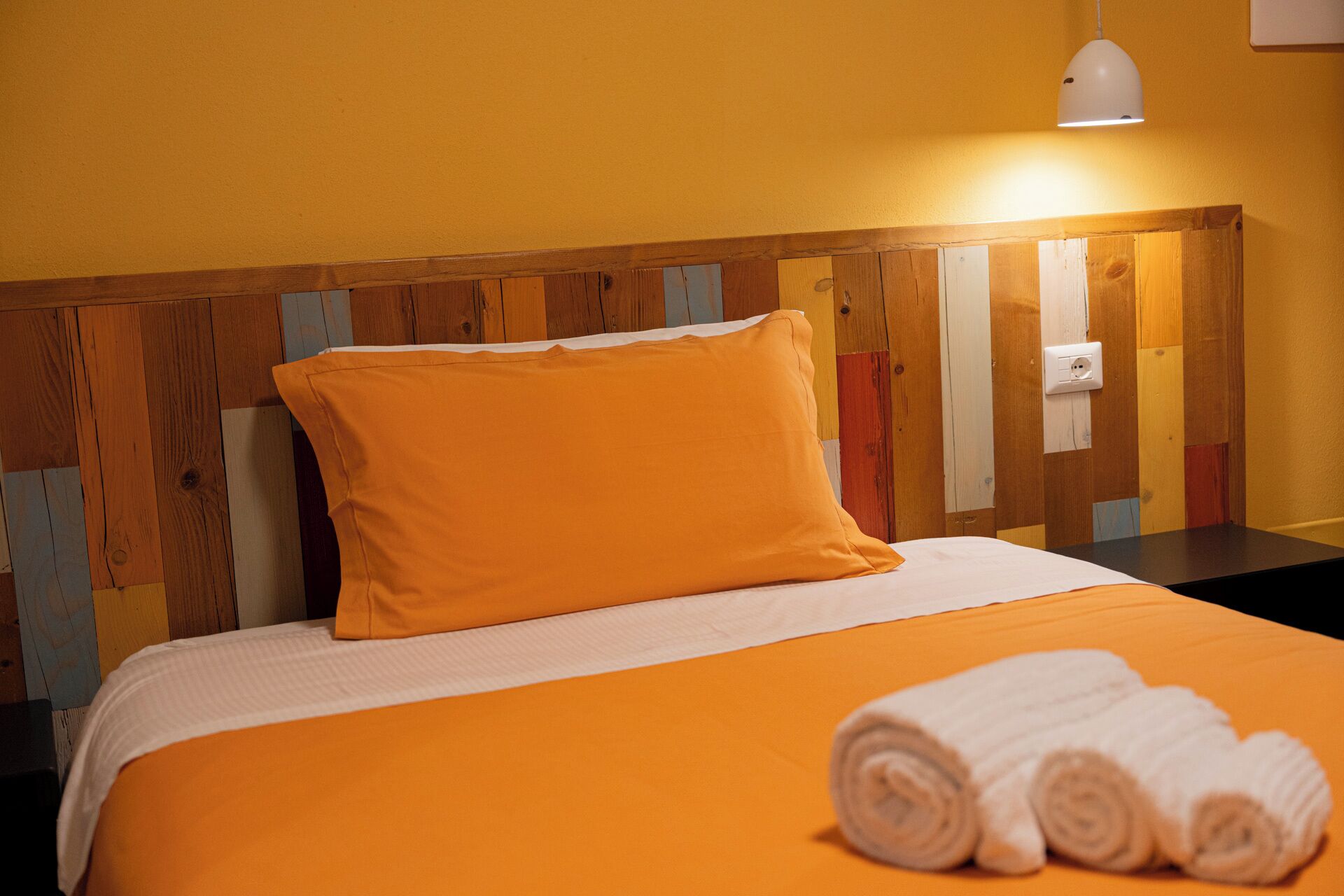 Quarto de hotel com cabeceira de madeira, colcha laranja, travesseiro branco e toalha branca enrolada na cama.
