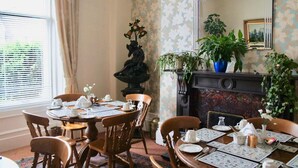 A dining room with a round table set for four, surrounded by wooden chairs. A fireplace with a mantel and potted plants is visible in the background. - Camillia Guest House (Aberdeen)