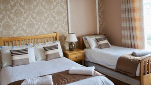 A hotel room with two single beds, a wooden headboard, a nightstand with a lamp, and a large window with curtains. - Camillia Guest House (Aberdeen)