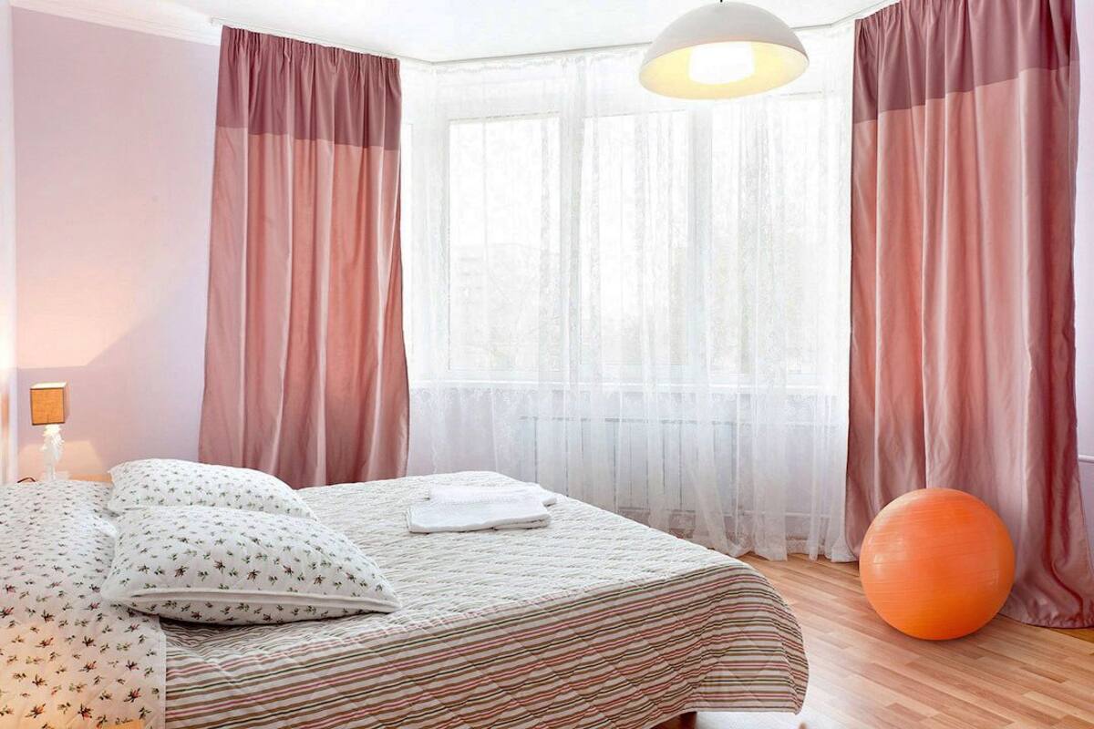 Ein Schlafzimmer mit einem Bett, einem Nachttisch, einer Lampe, einem Fenster mit durchsichtigen Vorhängen und einem Gymnastikball.
