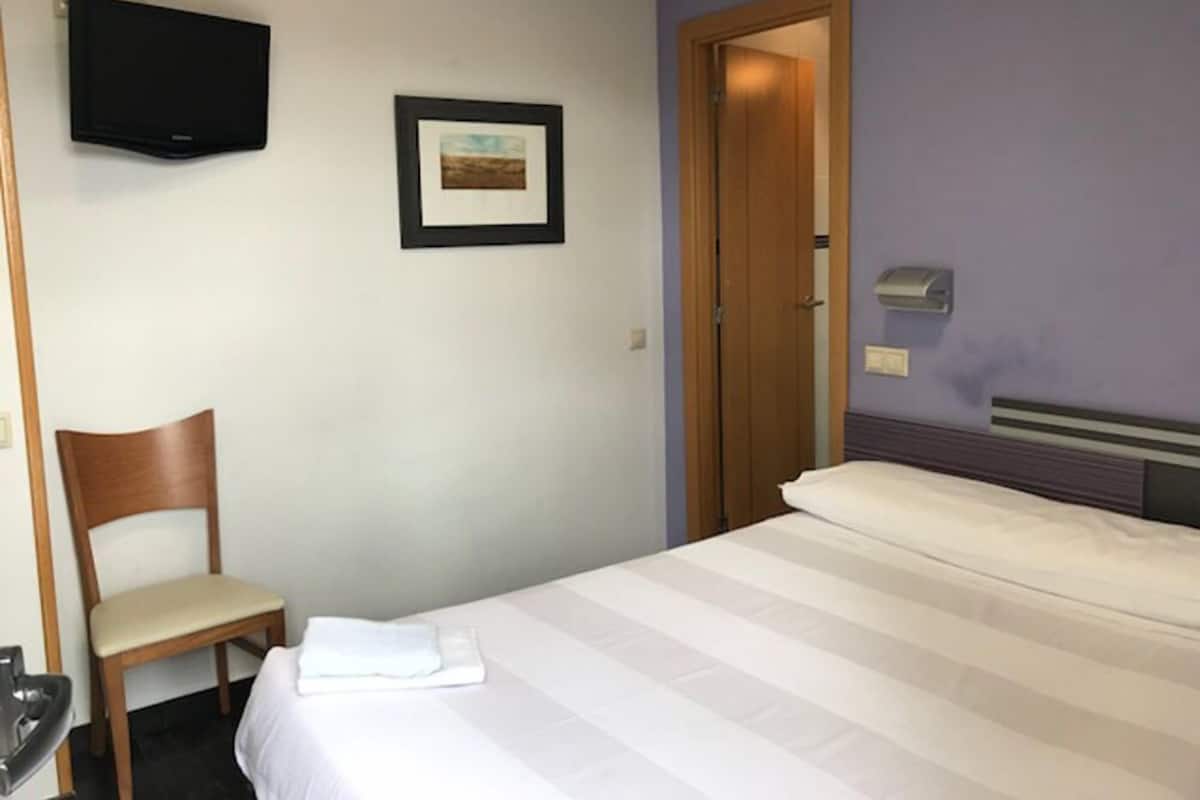 Hotellihuone, jossa on sänky, tuoli, seinälle asennettu televisio ja ovi.