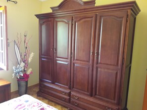 A bedroom with a large wooden wardrobe, a bed with a floral bedspread, and a vase with flowers.