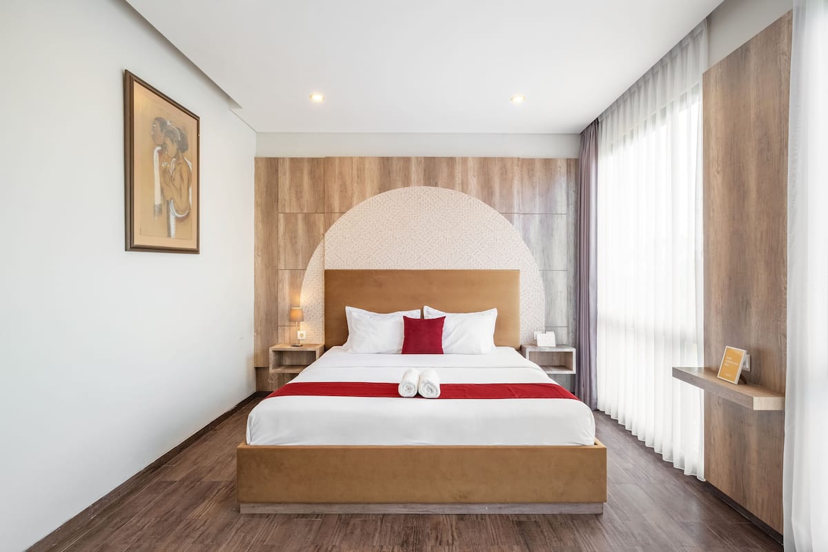 Una habitación de hotel moderna con una cama grande, cabecera de madera y un ventanal con cortinas translúcidas.