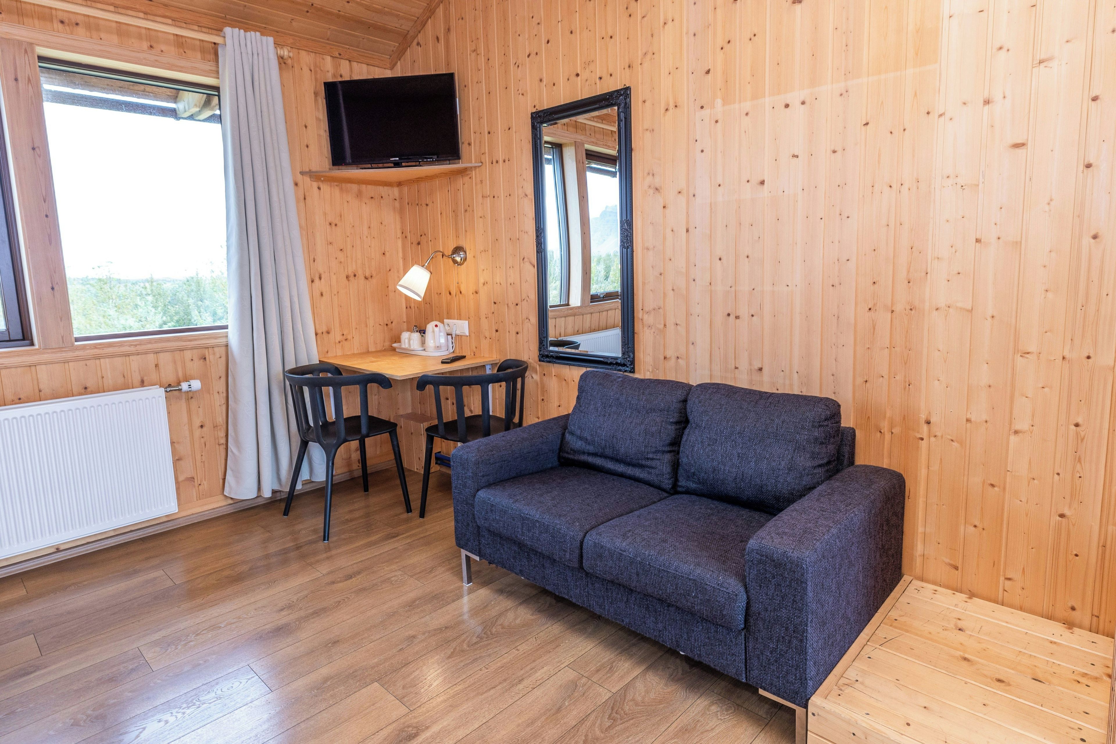 Una habitación con interior de madera, un sofá, una mesa con sillas, un televisor montado en la pared y una ventana con cortinas.