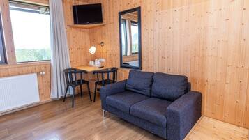 Una habitación con interior de madera, un sofá, una mesa con sillas, un televisor montado en la pared y una ventana con cortinas.