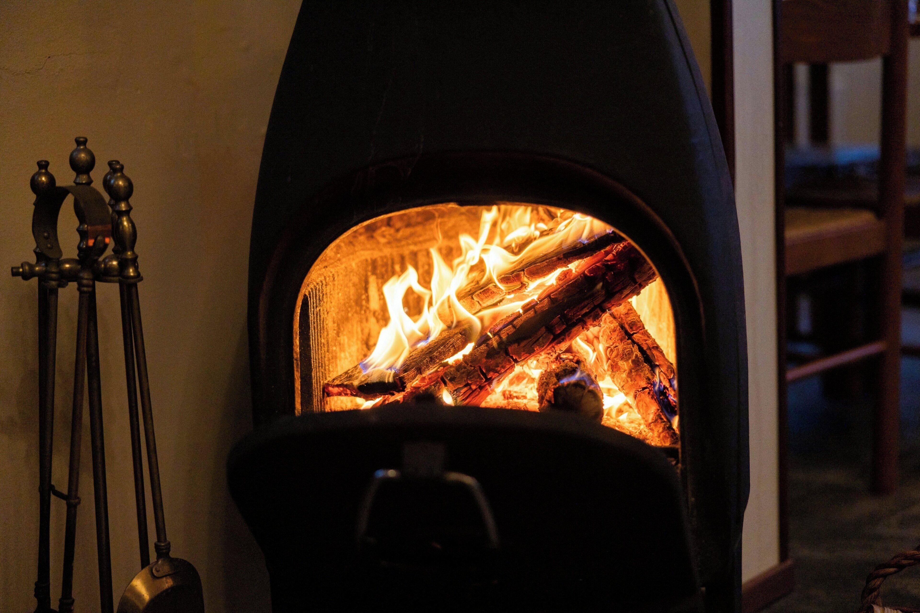 Ein Kaminofen mit brennendem Holz, ein Metallständer mit Haken und ein Stuhl im Hintergrund.