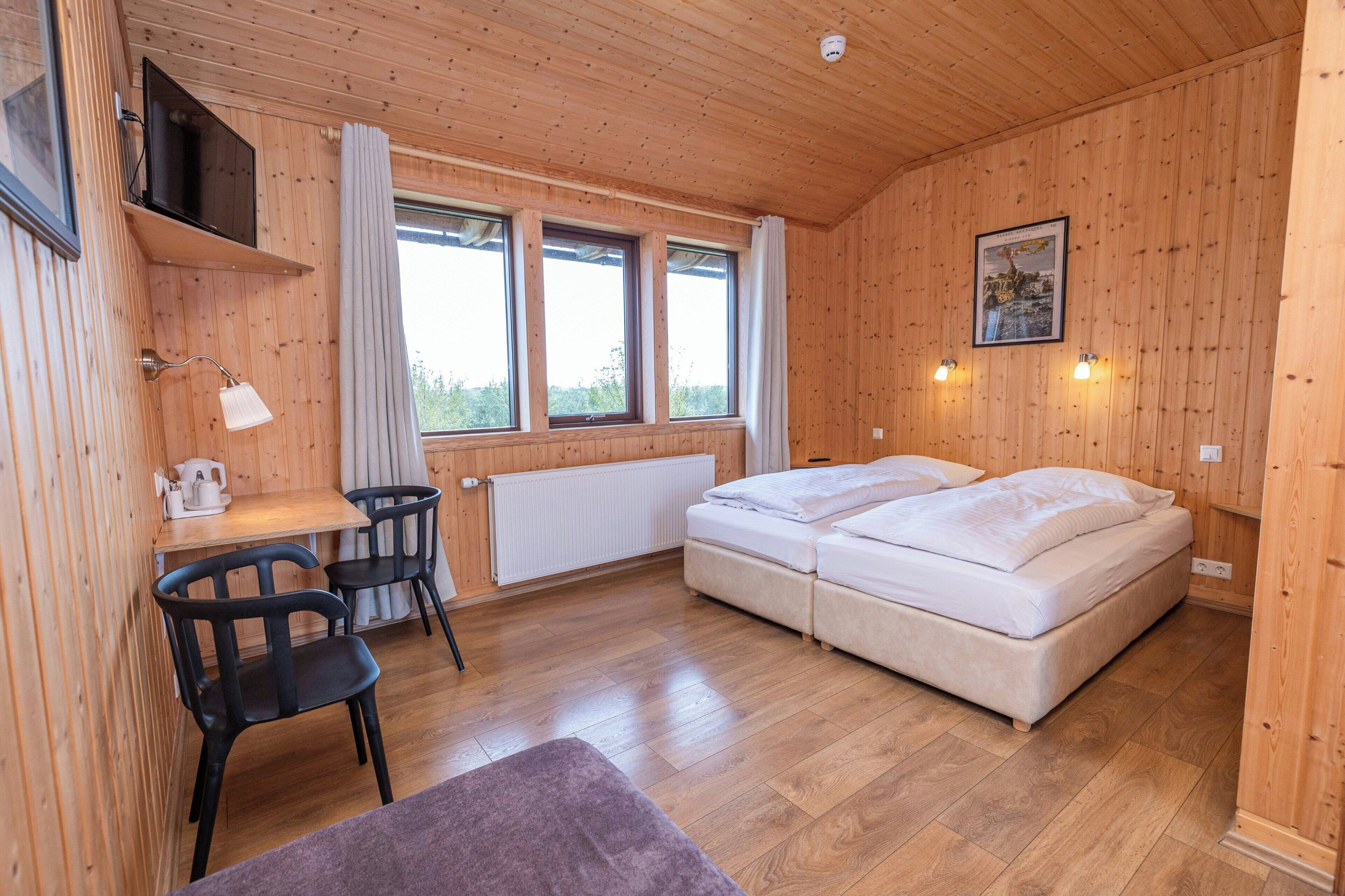 A wooden-paneled room with a bed, a desk, two chairs, and a radiator.