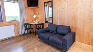 A room with a wooden floor, a sofa, a table with chairs, a TV mounted on the wall, and a window with curtains. - Arnanes Country Hotel (Höfn)