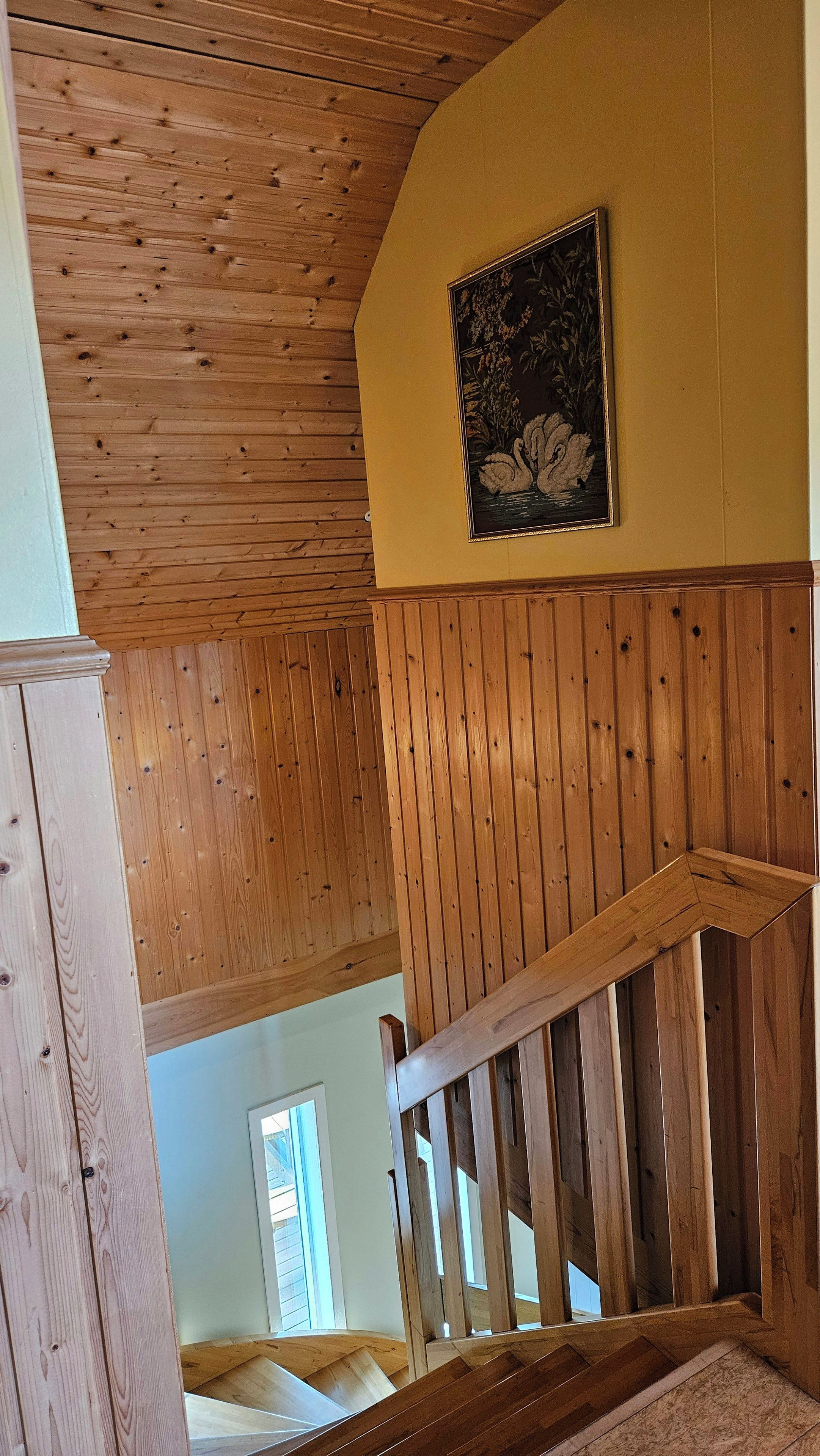 A wooden interior with a staircase, a framed picture on the wall, and a window in the background.