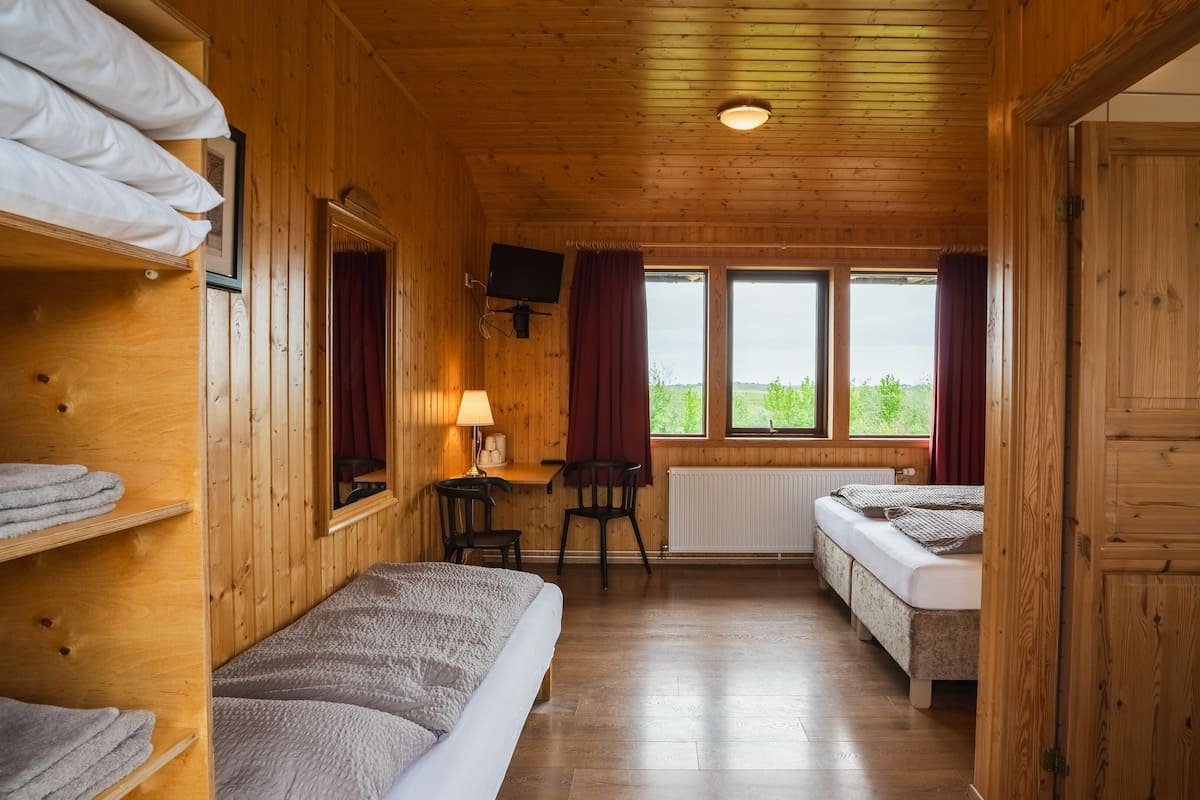 Quarto de cabana de madeira com beliches, uma escrivaninha com luminária e vista para o verde através de grandes janelas.