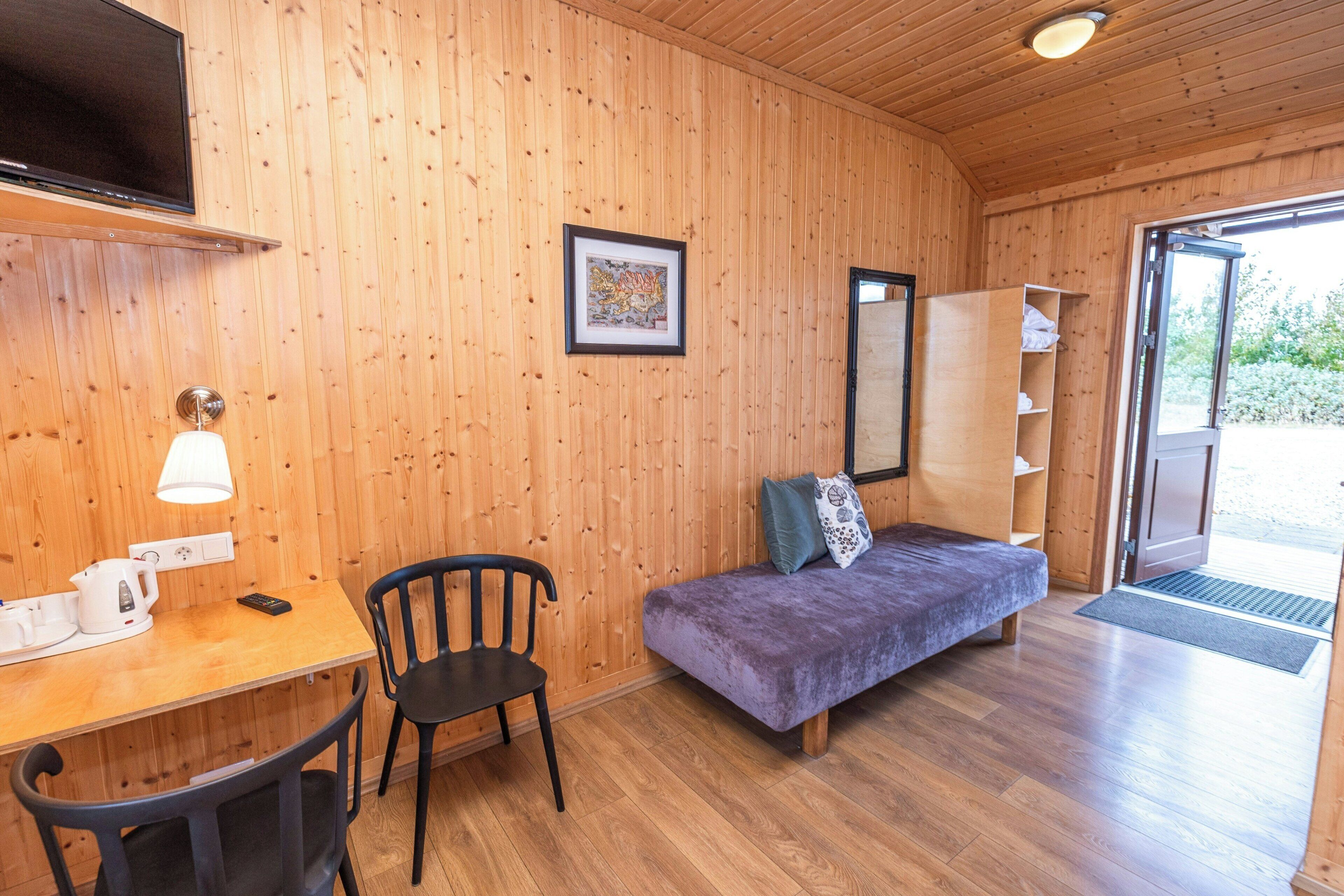 Ein Zimmer mit Holzwänden, einem Bett, einem Schreibtisch mit Lampe, einem Sessel und einem Fernseher.