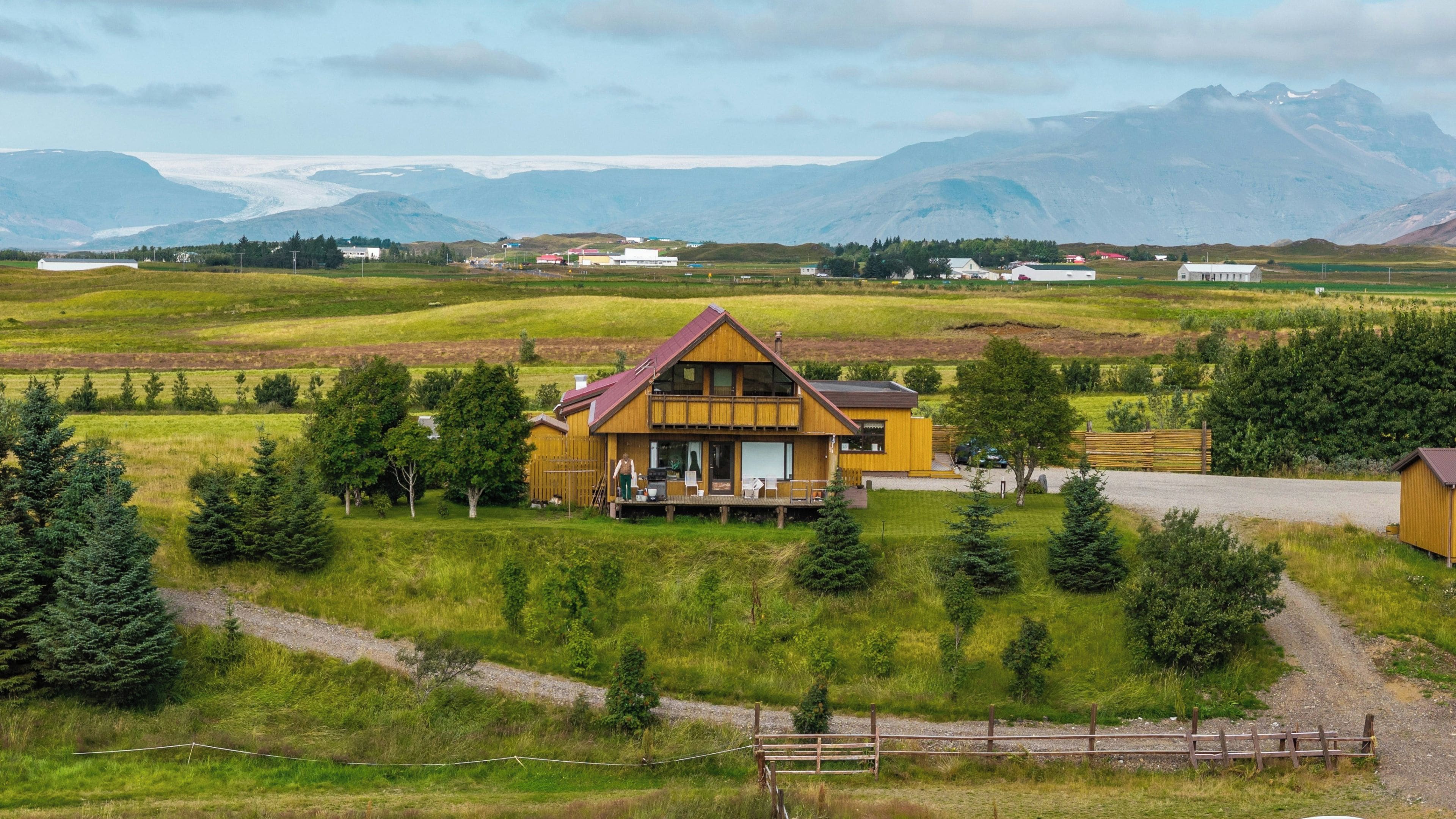 Casa de madera de dos pisos con balcón, rodeada de un paisaje rural con montañas al fondo.