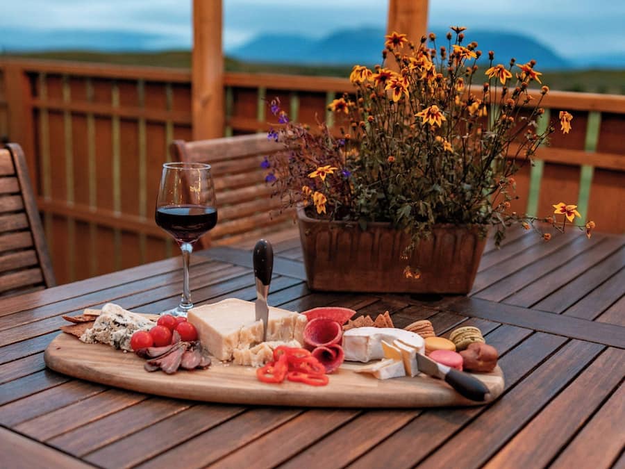 Une table en bois, une planche de charcuterie, un verre de vin rouge et une plante en pot aux fleurs jaunes.