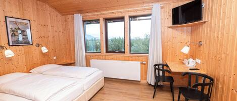 A wooden-paneled room with two beds, a desk, chairs, a radiator, and a TV mounted on the wall.