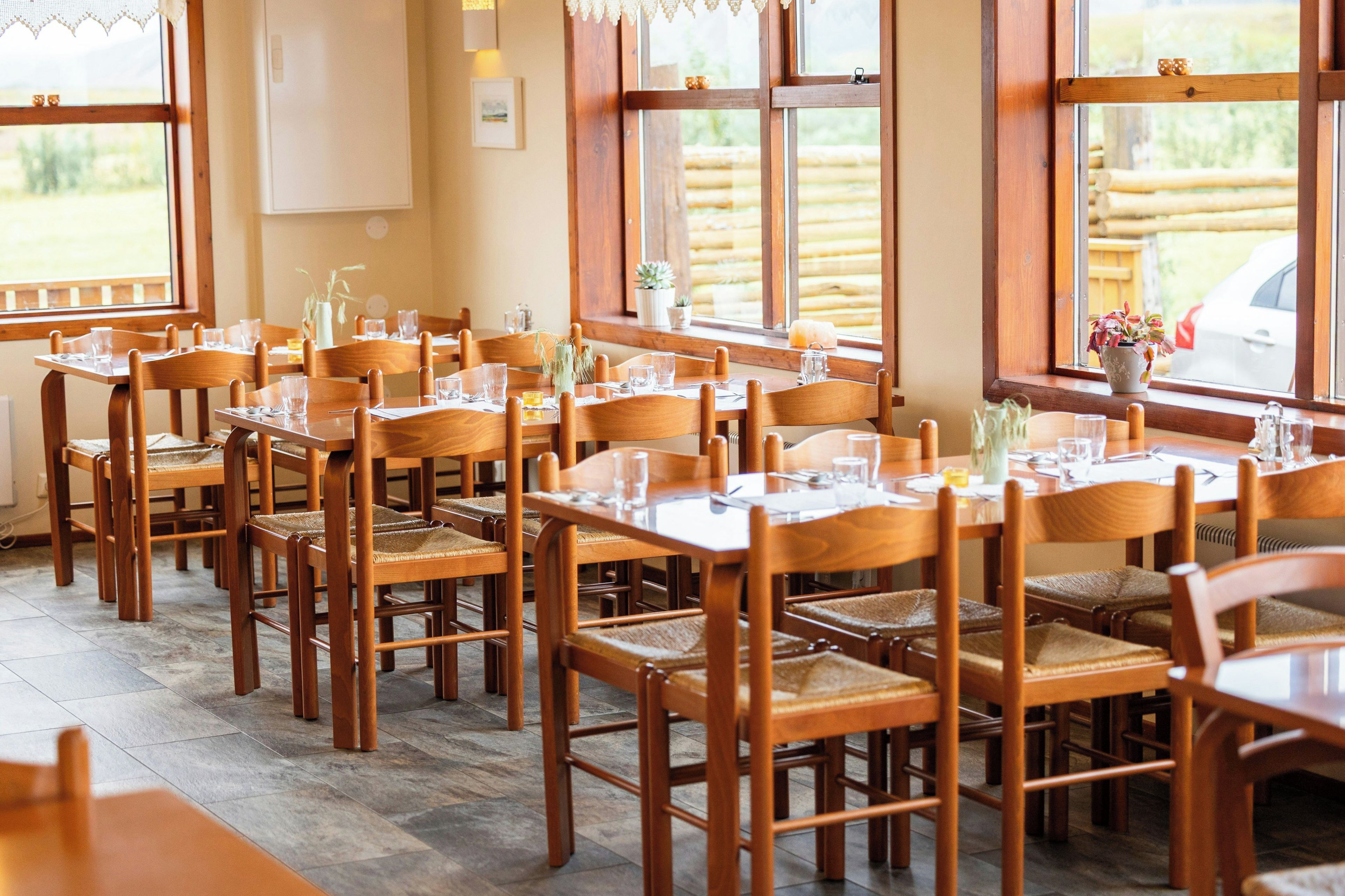 A restaurant with wooden tables and chairs, large windows, and a view of the outdoors.