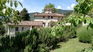 A stone building with a tiled roof, surrounded by greenery and hills in the background.