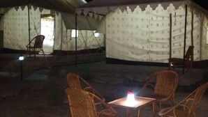 Un terrain de camping avec des tentes et des chaises en osier disposées autour d’une table centrale, le tout éclairé par une source de lumière.