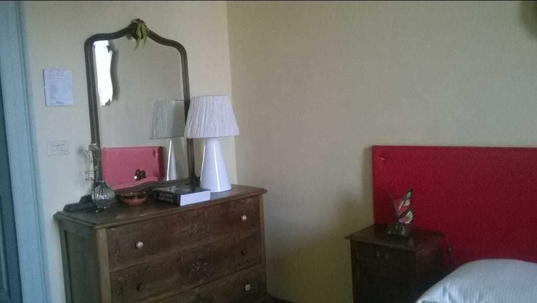 A bedroom with a wooden dresser, a mirror, a lamp, a red headboard, and a bedside table with a decorative item.