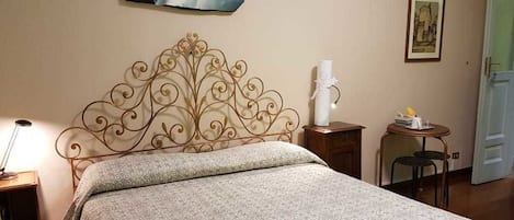Una cama bien hecha con una colcha estampada, un cabecero de metal decorativo, una mesita de noche con una lámpara y un cuadro en la pared.