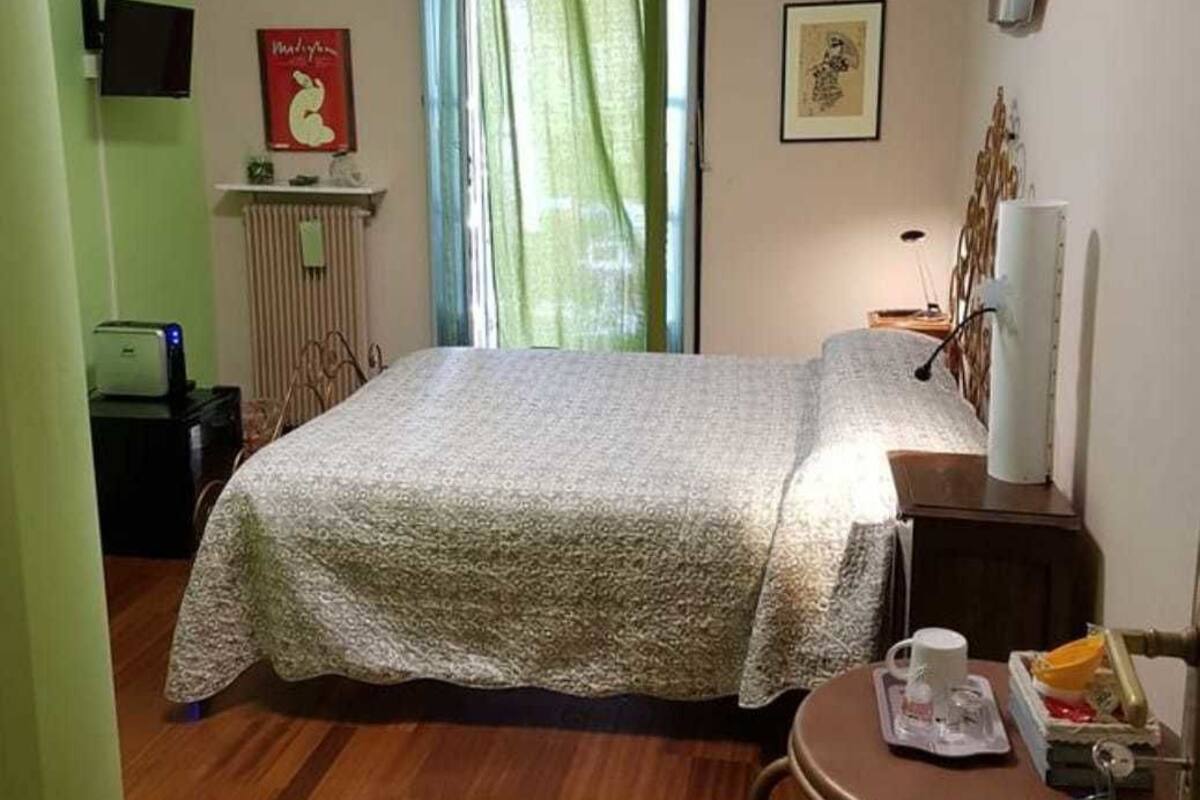 A bedroom with a bed, a small table with a cup and a book, a chair, a radiator, and a window with a curtain.