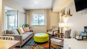 A modern living room with a sofa, ottoman, chairs, a dining table, and a TV mounted on the wall. - WoodSpring Suites Naples East (Naples)