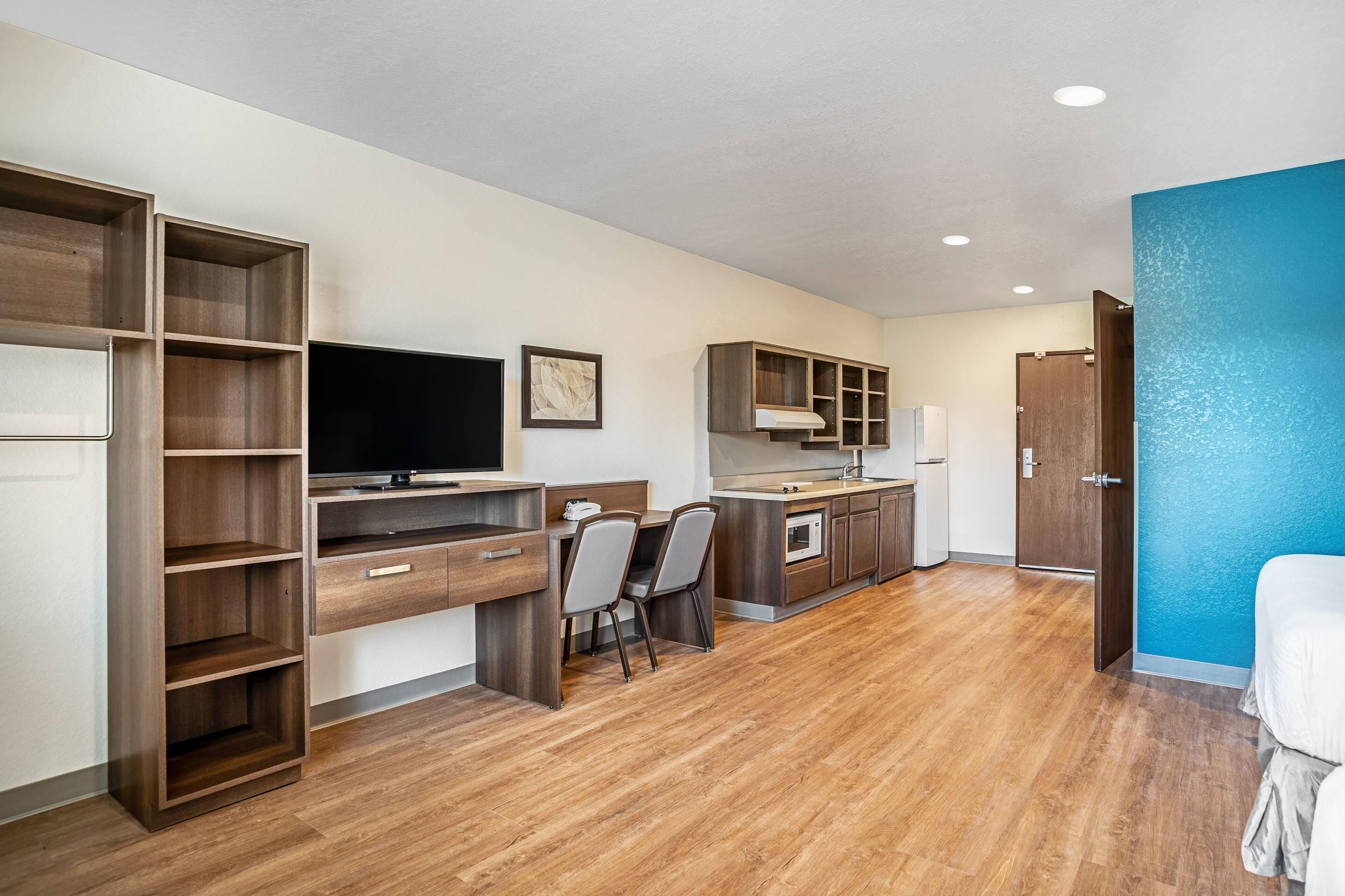 A modern kitchen with wooden cabinets, a dining area with chairs, a TV, and a blue accent wall.