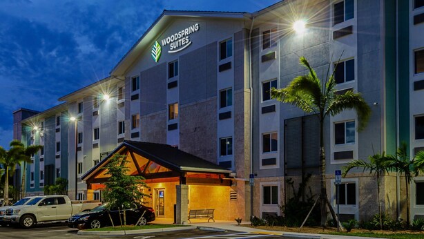 A hotel with a sign reading "Woodspring Suites." It has multiple floors, a covered entrance, and palm trees in the foreground. - WoodSpring Suites Naples East (Naples)