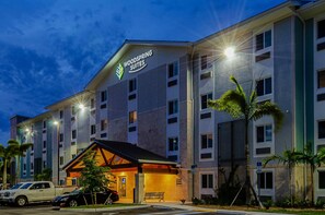 A hotel with a sign reading "Woodspring Suites." It has multiple floors, a covered entrance, and palm trees in the foreground.