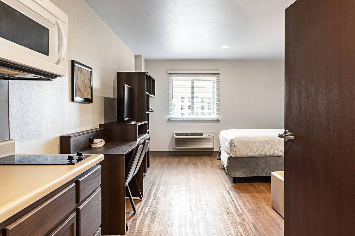 Une chambre à coucher comprenant un lit, un bureau, une chaise, une commode, un four à micro-ondes, un tableau encadré et une fenêtre avec des stores.