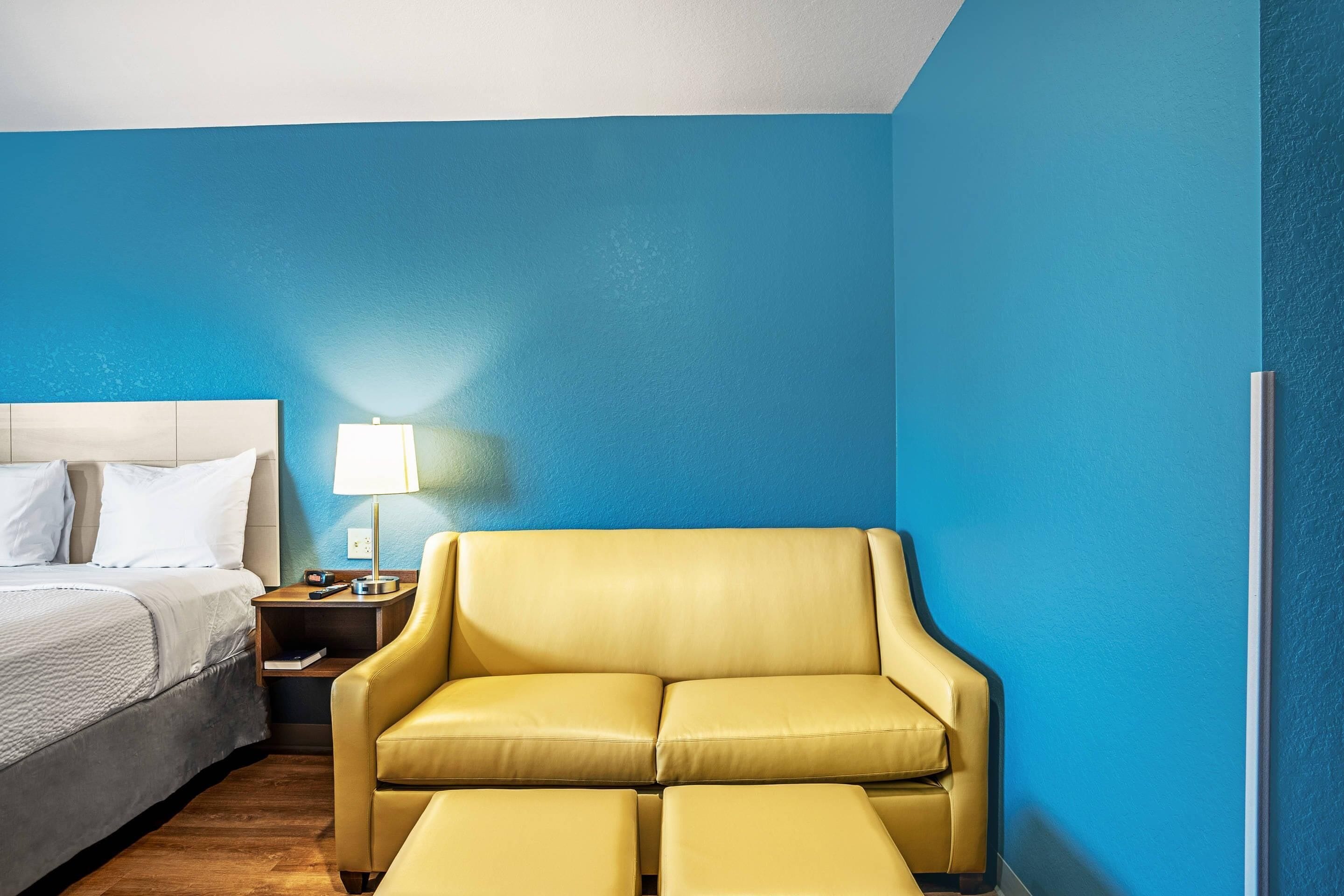 A hotel room with a bed, a yellow leather sofa, a nightstand with a lamp, and a blue wall.