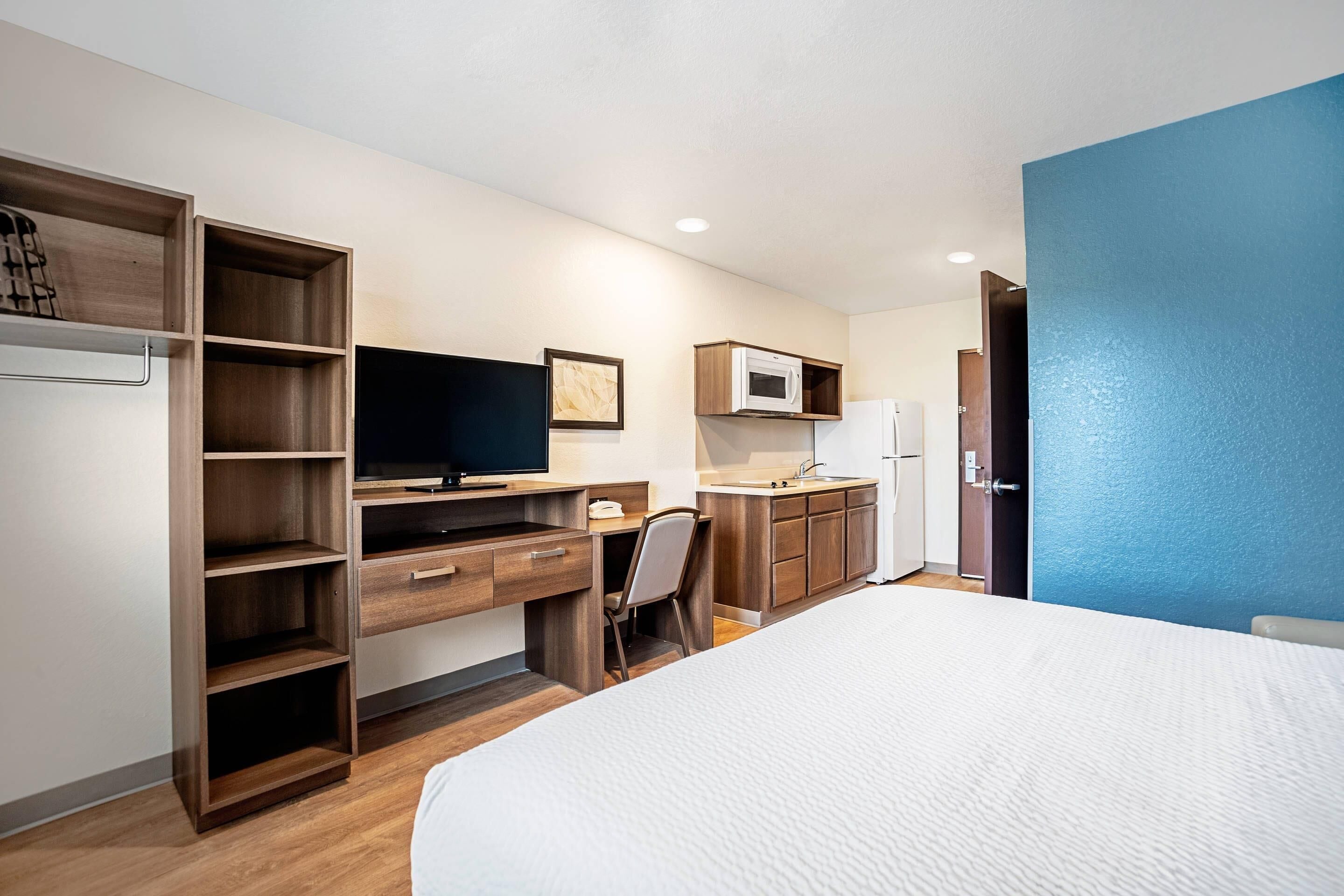 A hotel room with a bed, a desk with a computer, a chair, a microwave, and a refrigerator.