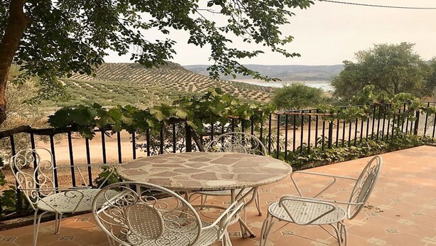 Un patio con una mesa redonda y sillas, rodeado por una valla de metal y vistas a viñedos y colinas.