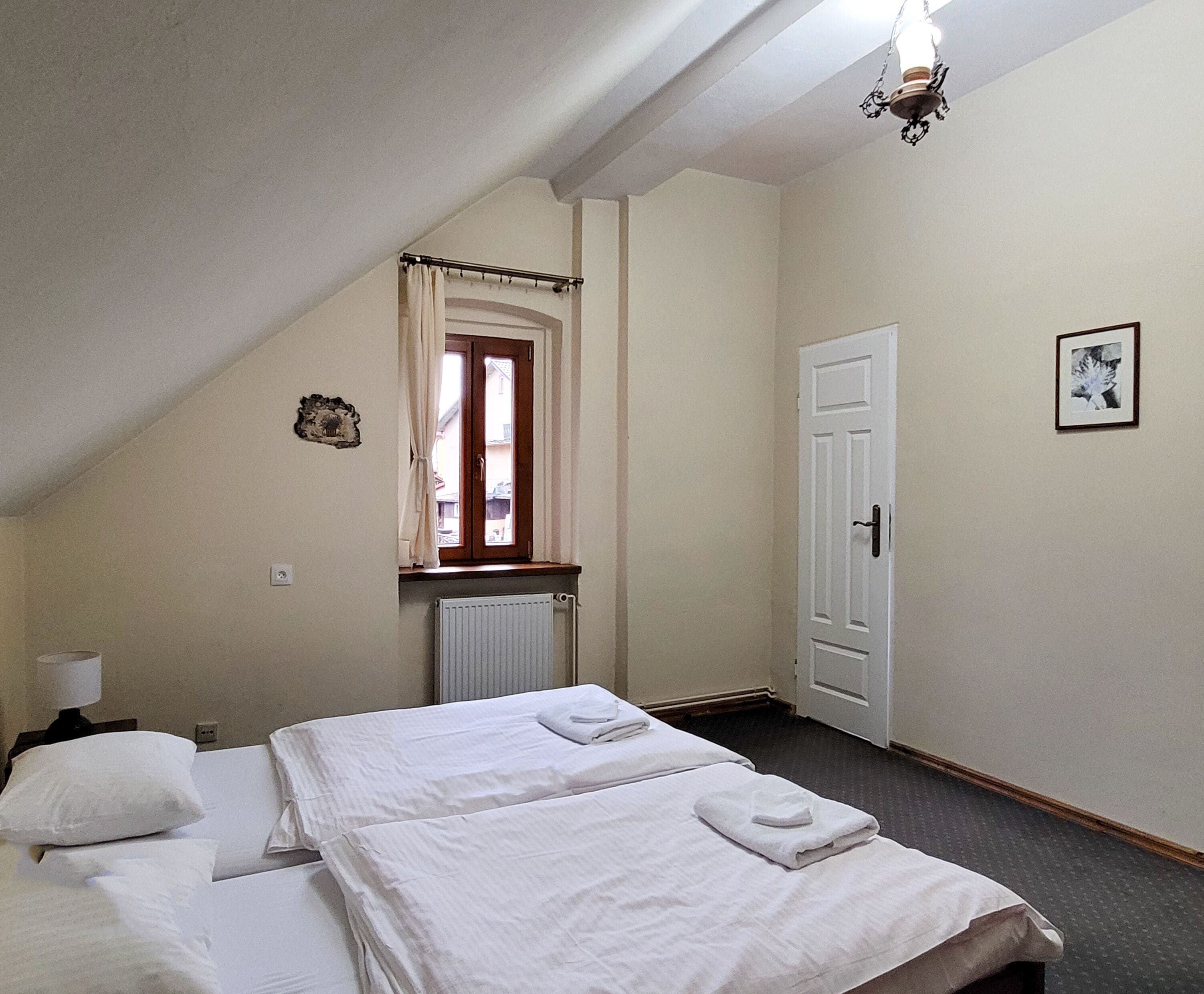 A bedroom with two beds, a window with curtains, a radiator, a door, and a picture on the wall.